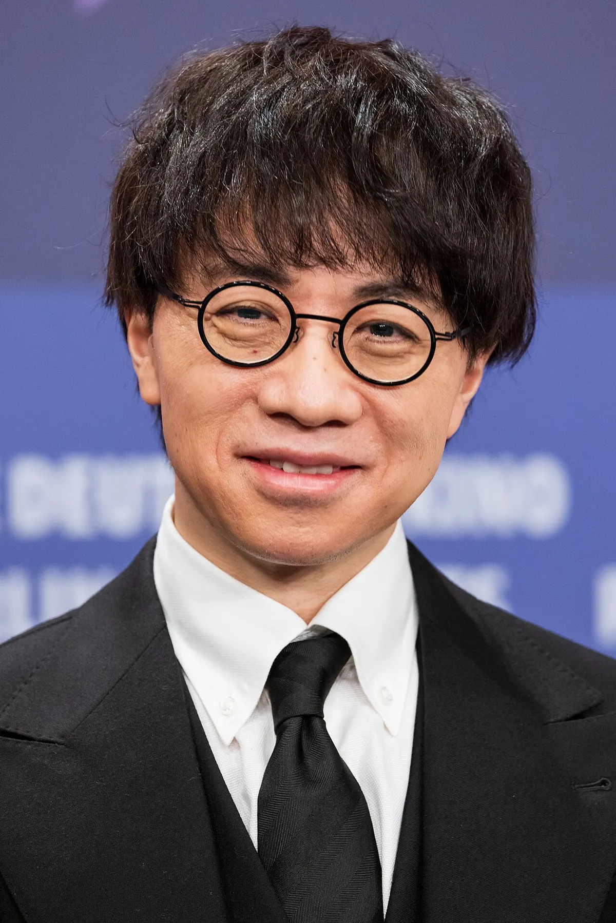 Makoto Shinkai berkacamata bulat, berjas hitam dan berdasi, tersenyum di depan latar biru bertuliskan Berlinale