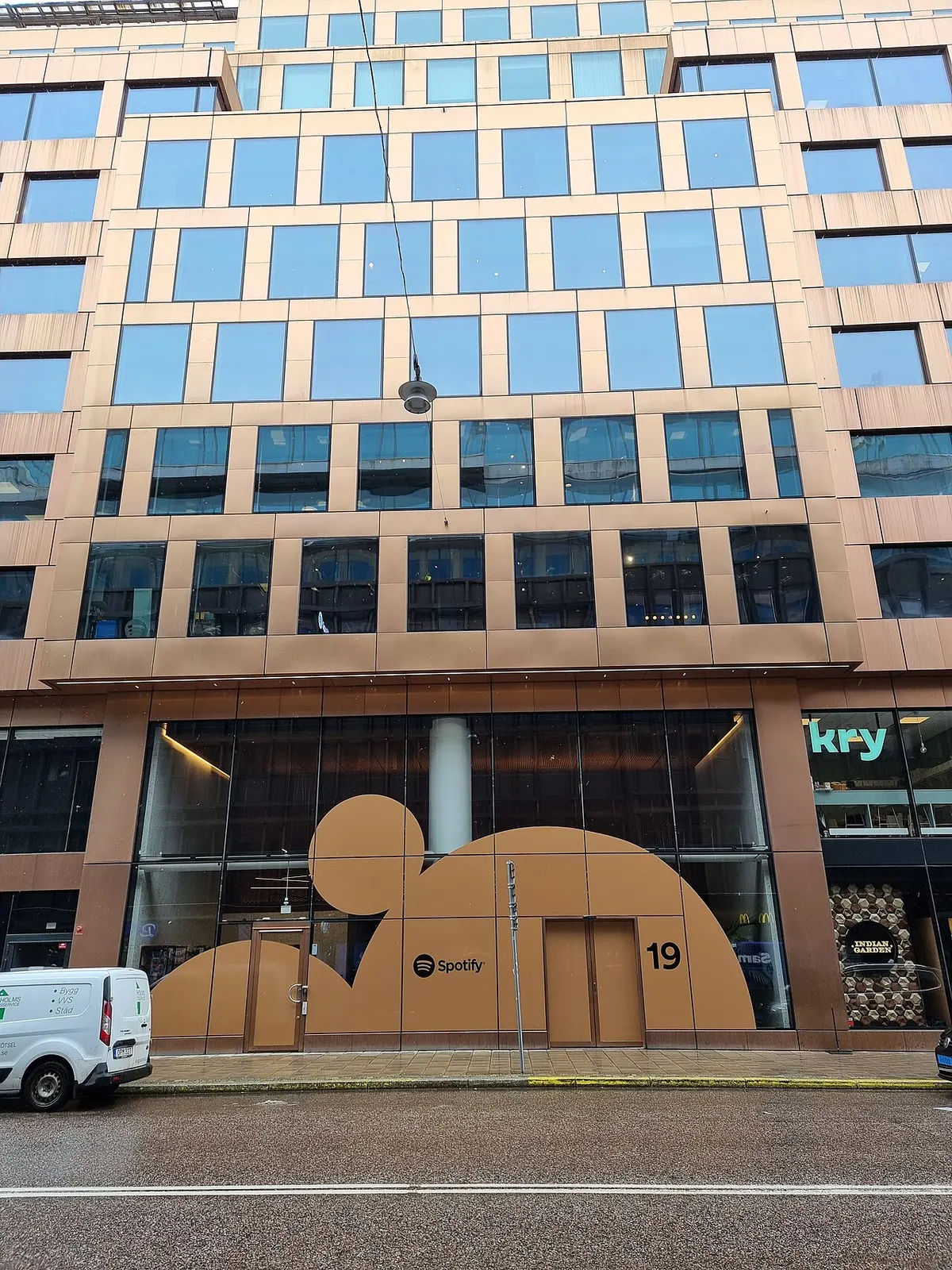 Fasad gedung kantor berjendela kaca dengan bingkai tembaga, dengan logo Spotify dan angka 19 di kaca lantai dasar, di tepi jalan Stockholm