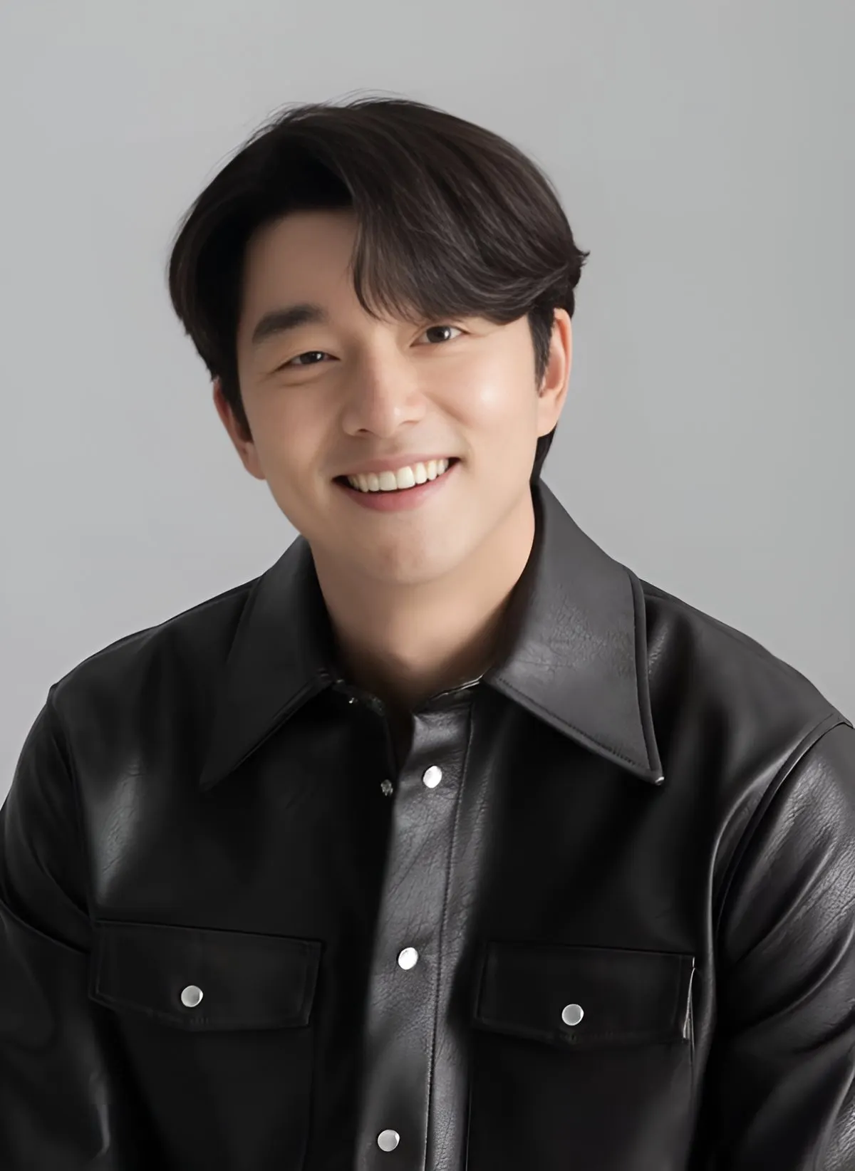 Gong Yoo berkemeja kulit hitam tersenyum lebar di depan latar abu-abu polos
