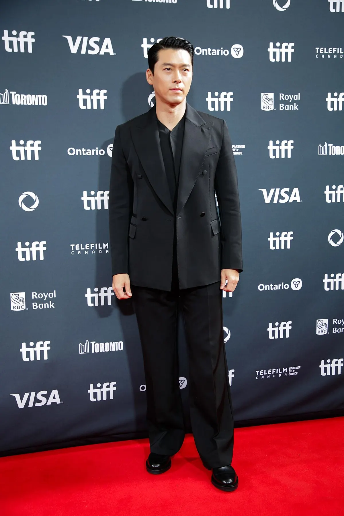 Hyun Bin berjas hitam berdiri di karpet merah dengan latar papan berlogo tiff dan sponsor