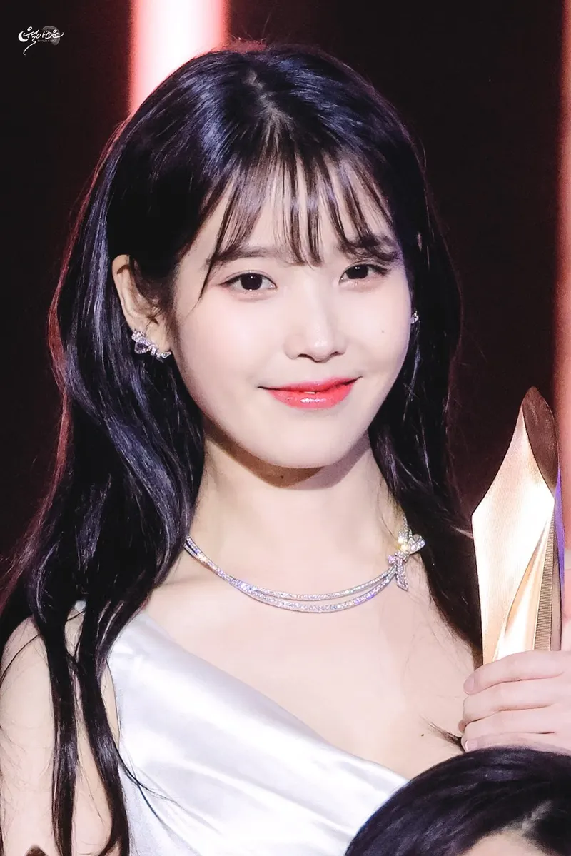 IU berambut panjang berponi dan bergaun putih tersenyum sambil memegang piala berwarna keemasan