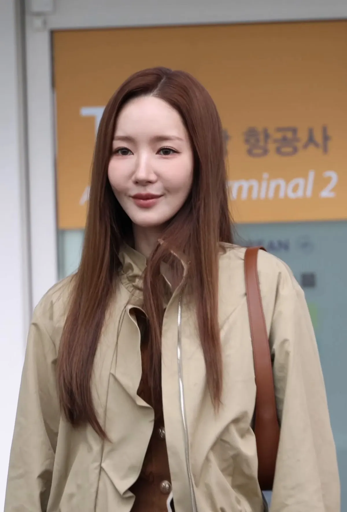 Park Min-young berambut panjang cokelat dan berjaket krem berdiri di depan papan petunjuk Terminal 2 bandara