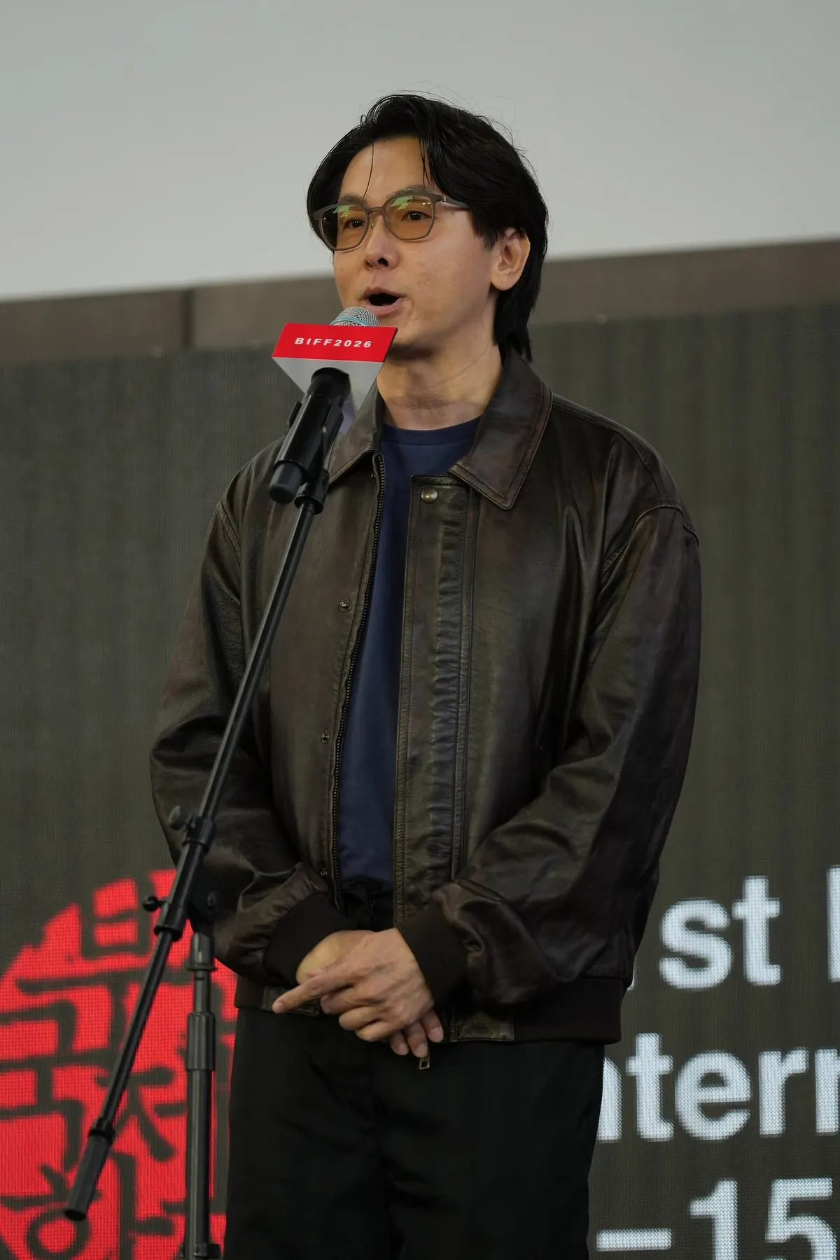Banjong Pisanthanakun berkacamata dan berjaket kulit cokelat berbicara di depan mikrofon berlabel BIFF2026