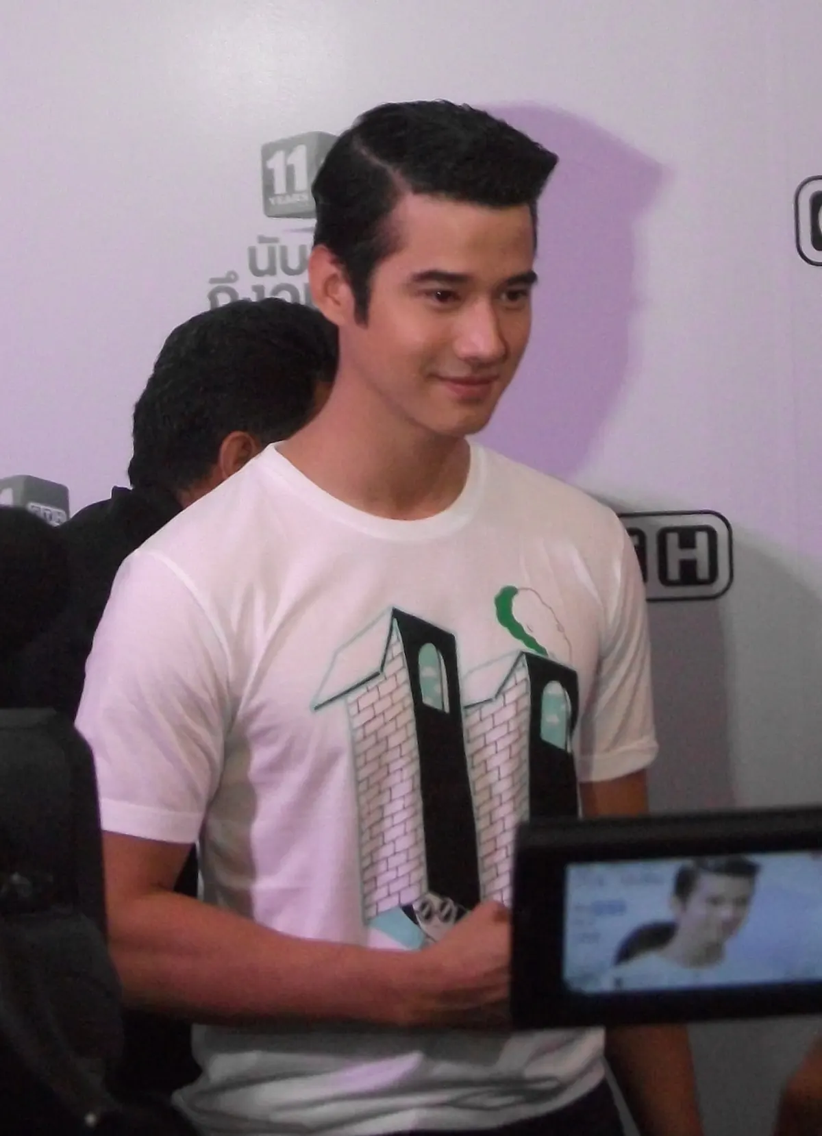 Mario Maurer berkaus putih bergambar angka 11 tersenyum di depan dinding acara, dengan layar kamera wartawan di sudut kanan bawah