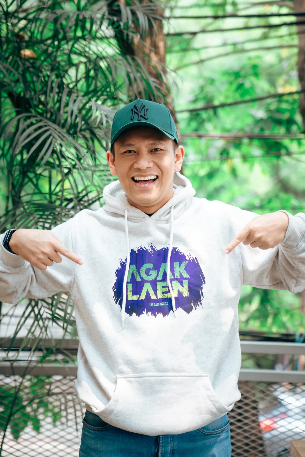 Boris Bokir bertopi hijau tersenyum lebar sambil menunjuk logo Agak Laen di hoodie abu-abunya