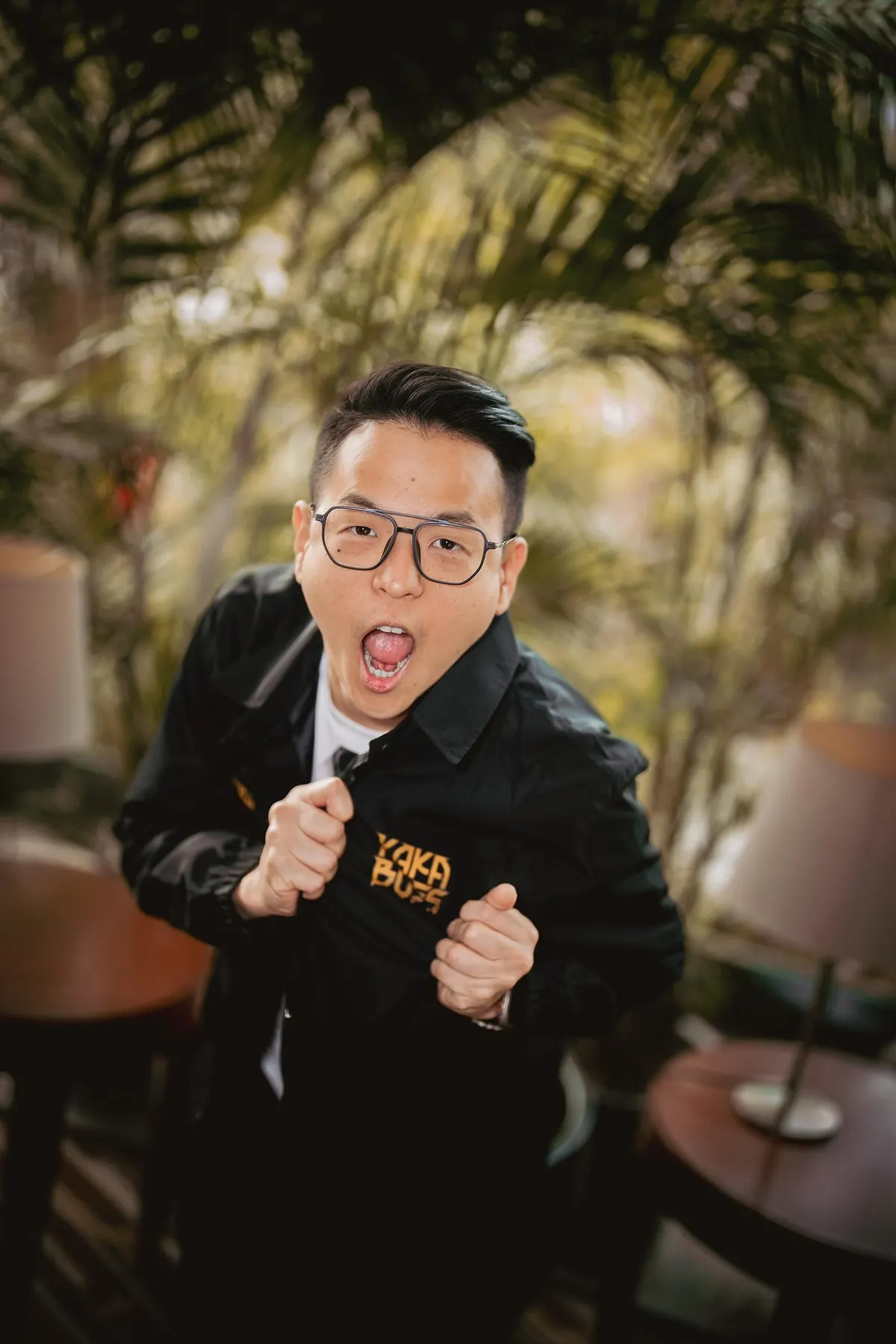 Ernest Prakasa berkacamata dan berjaket hitam berpose dengan mulut terbuka di depan dedaunan palem