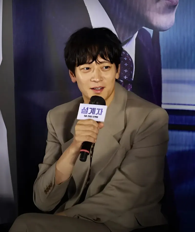 Gang Dong-won berjas krem duduk sambil berbicara memegang mikrofon berlabel judul film di depan poster