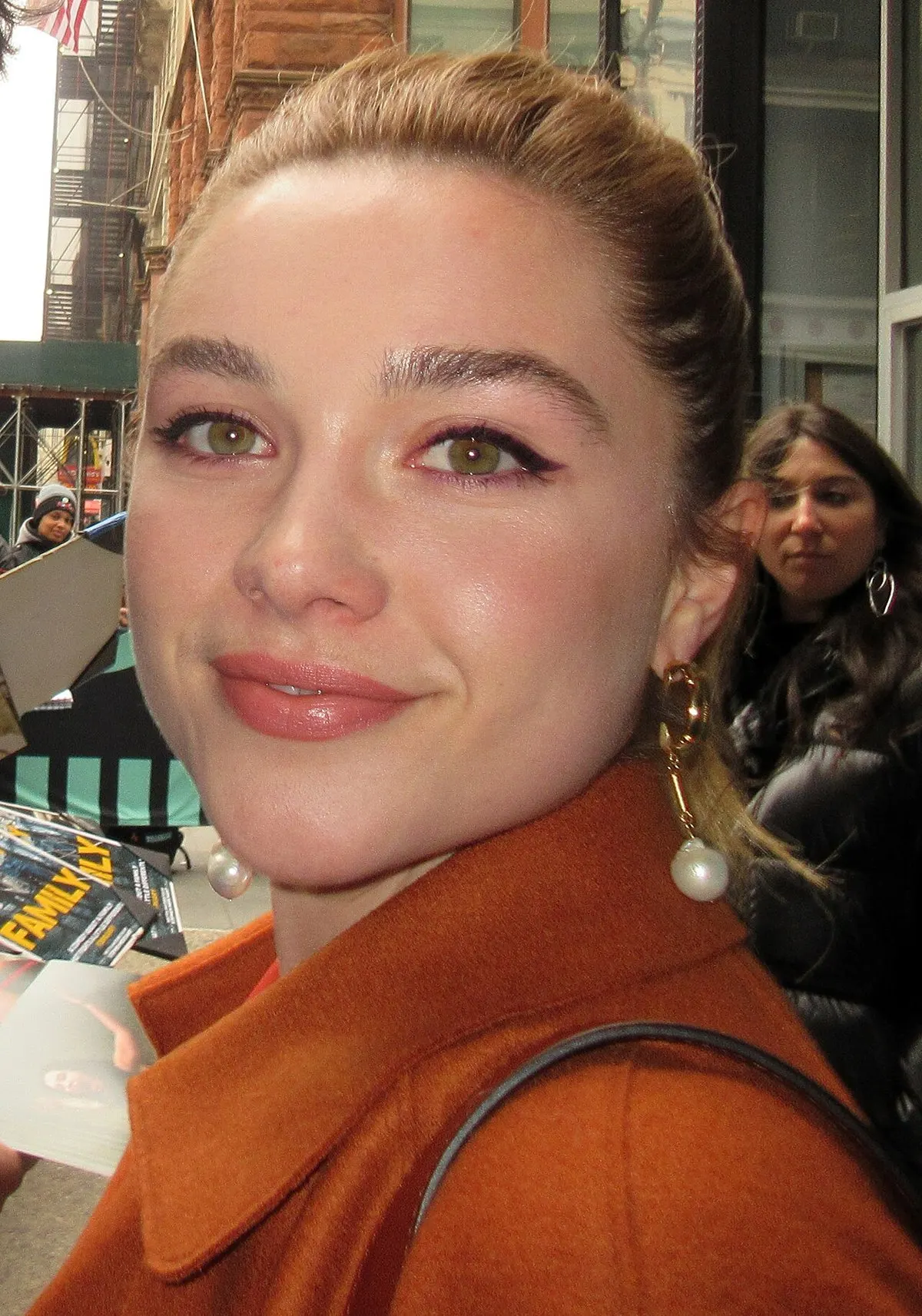 Florence Pugh berambut diikat dan bermantel oranye tersenyum ke arah kamera di tepi jalan kota pada 2019