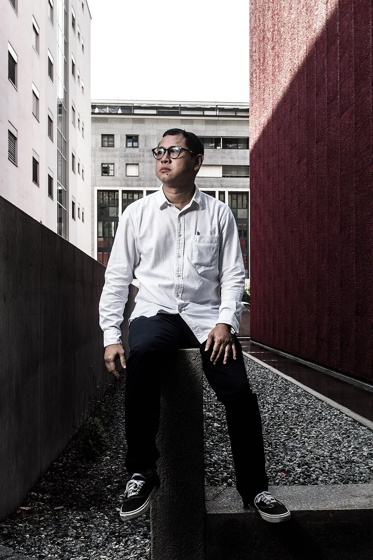 Sutradara Edwin berkemeja putih dan berkacamata duduk di tembok rendah di antara gedung saat Festival Film Locarno 2021