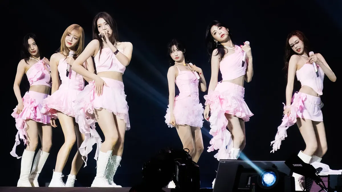 Anggota BABYMONSTER berbusana merah muda berumbai tampil berjajar di panggung konser berlatar gelap dengan lampu sorot biru