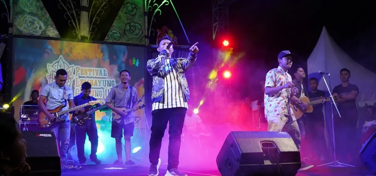 Denny Caknan berjaket motif bernyanyi sambil menunjuk ke penonton, diapit para pemain gitar di panggung Festival Lembayung Gunung Ukir
