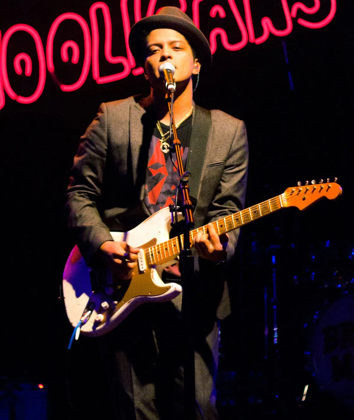 Bruno Mars bertopi fedora dan berjas abu-abu bernyanyi sambil bermain gitar listrik putih di depan tulisan neon merah Hooligans