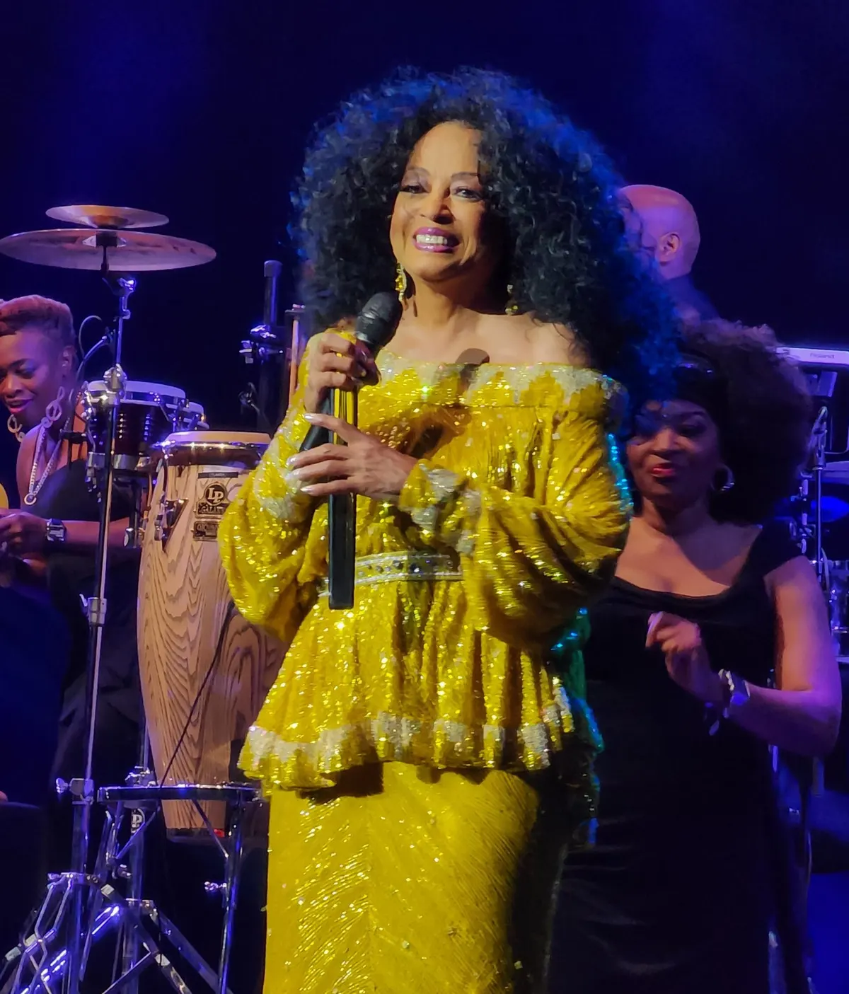 Diana Ross bergaun kuning berpayet tersenyum sambil memegang mikrofon di panggung, dengan pemain perkusi dan penyanyi latar di belakangnya