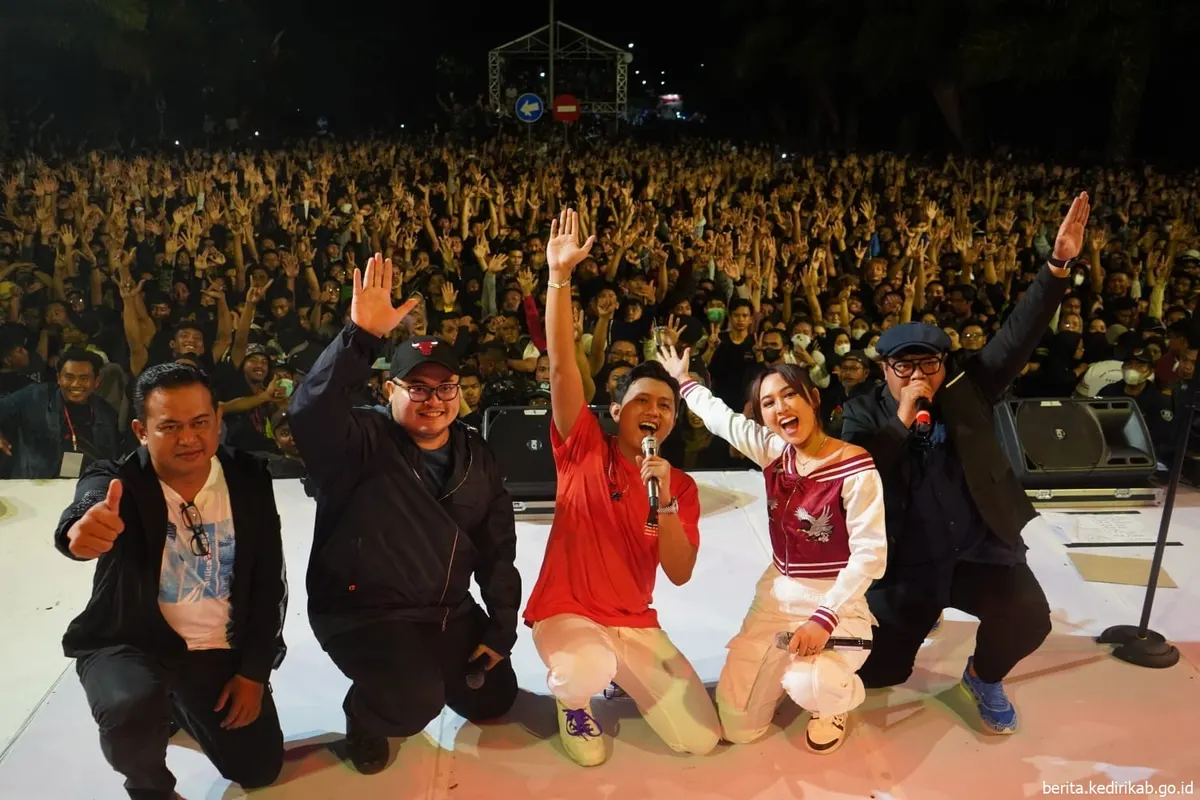 Denny Caknan berkaus merah dan Happy Asmara berjaket varsity merah putih berlutut di panggung sambil melambai ke ribuan penonton, bersama tiga pria lain