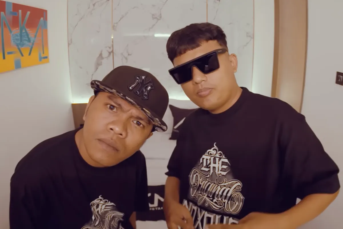 Dua personel NDX A.K.A. berkaus hitam bergambar tulisan The Original, satu bertopi dan satu berkacamata hitam, menatap kamera di dalam ruangan