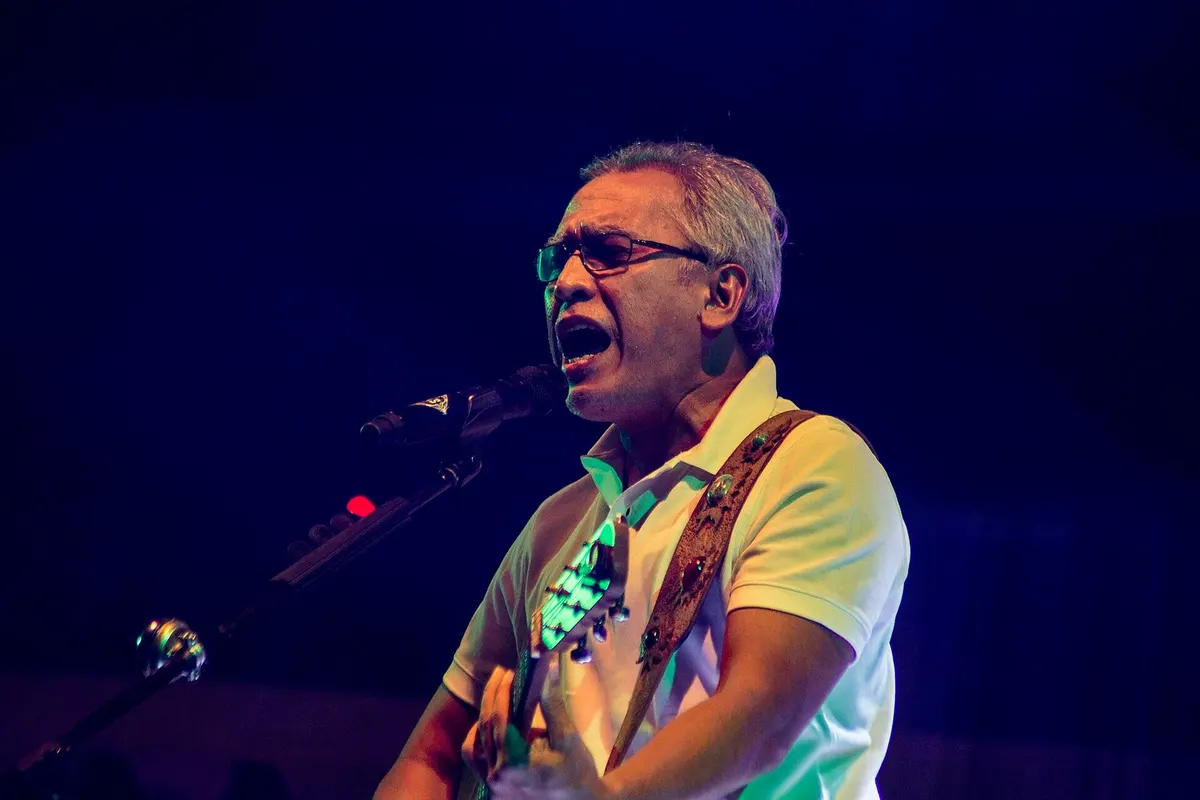 Iwan Fals berkacamata dan berkaus polo putih bernyanyi di depan mikrofon sambil memainkan gitar di panggung bercahaya biru