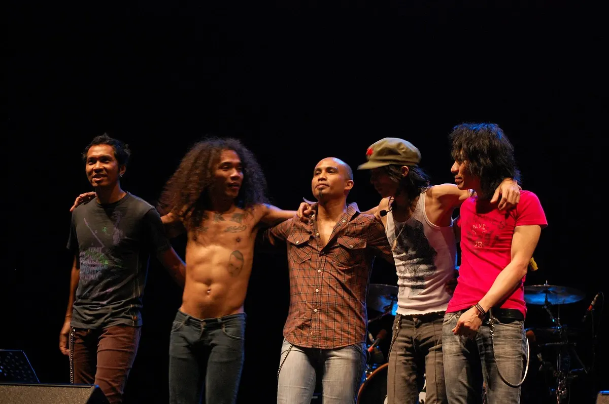 Lima personel Slank berdiri berangkulan di panggung berlatar gelap, salah satunya bertopi hijau dan satu lagi bertelanjang dada