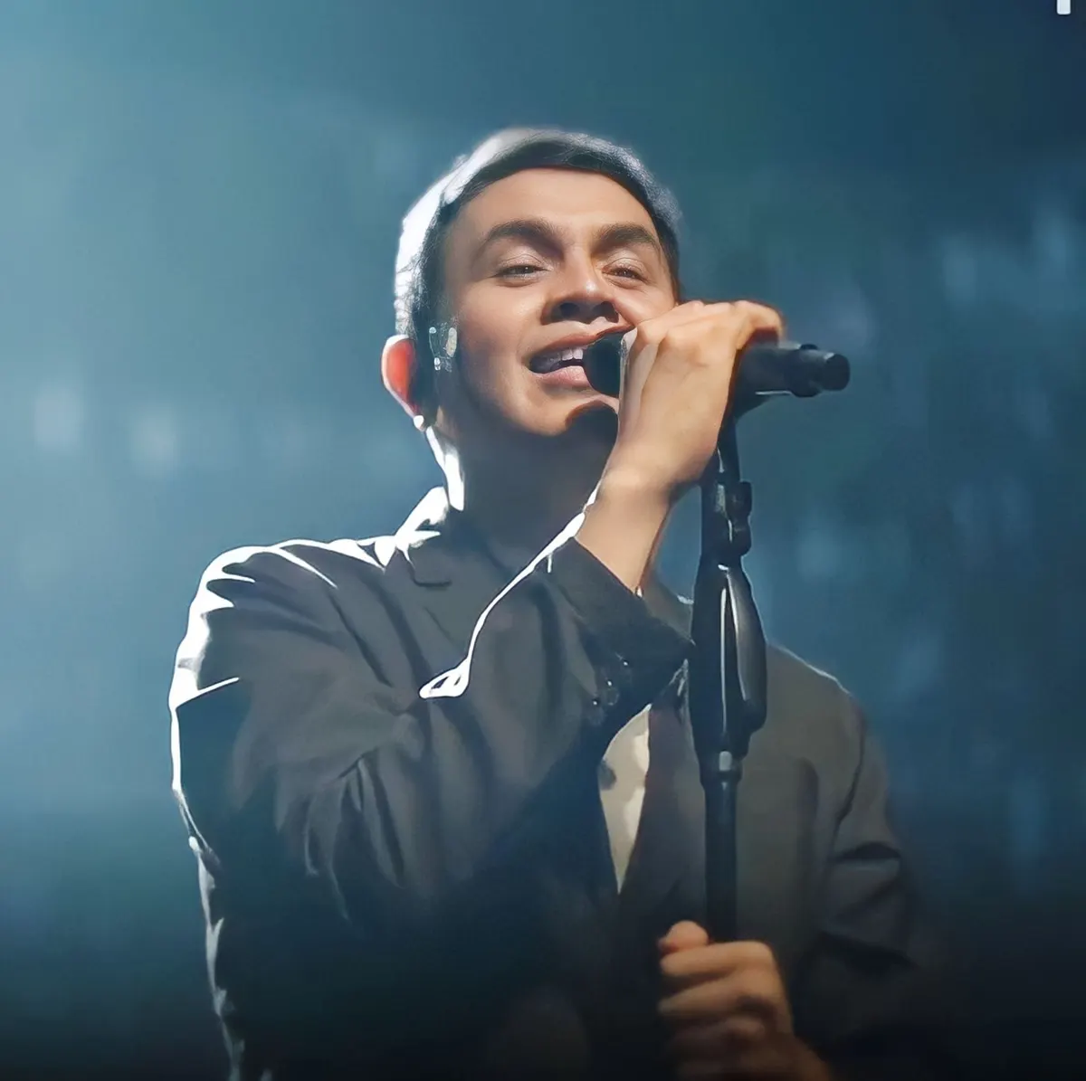 Tulus berjas gelap bernyanyi sambil memegang mikrofon di stand, dengan latar panggung biru berkabut