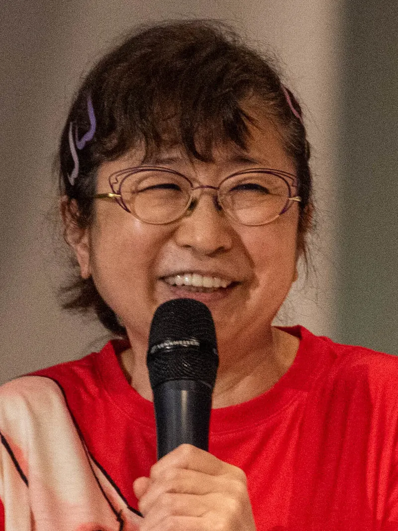 Mayumi Tanaka berkacamata dan berkaus merah tertawa sambil memegang mikrofon di atas panggung