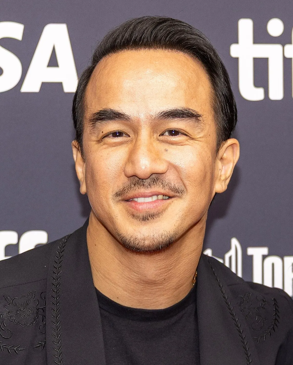 Joe Taslim berjas hitam bersulam dan berkaus hitam tersenyum di depan latar logo Toronto International Film Festival 2025
