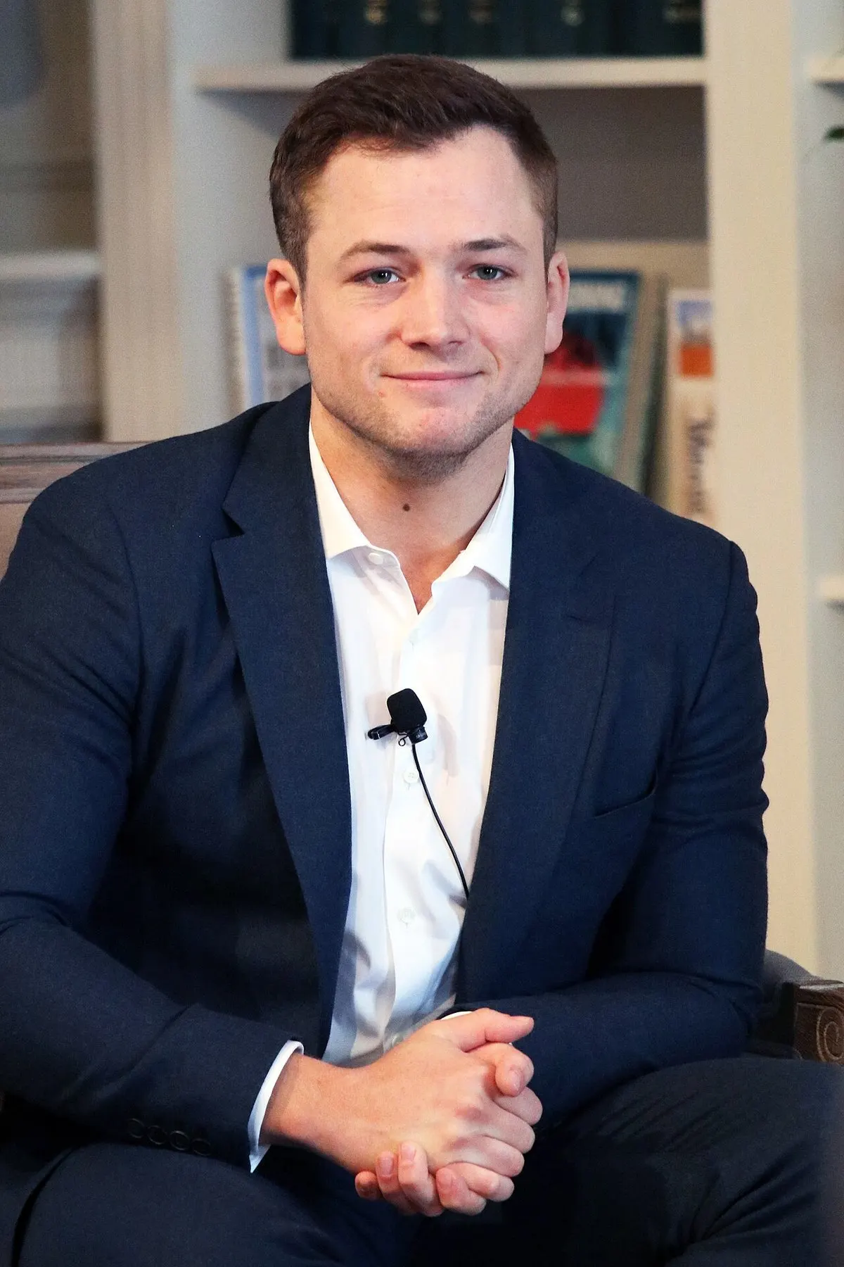 Taron Egerton berjas biru tua dan kemeja putih dengan mikrofon klip duduk di kursi di depan rak buku