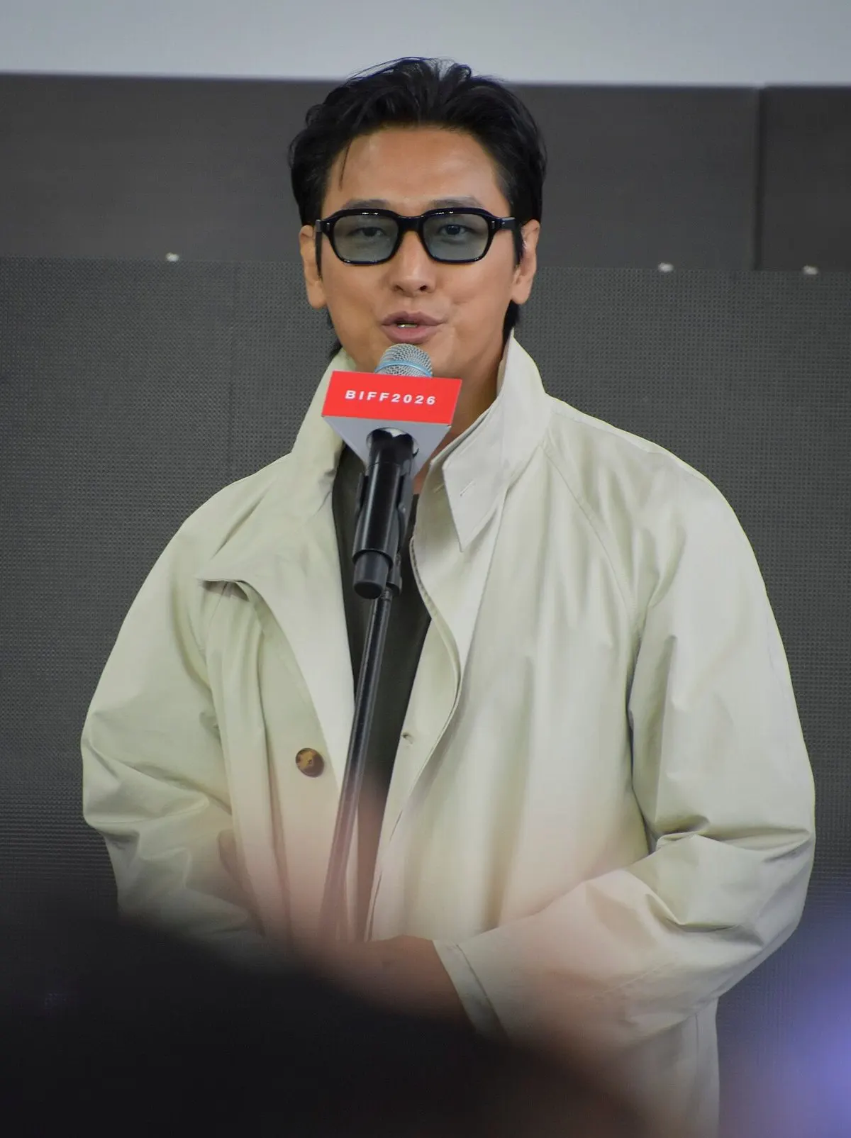 Ju Ji-hoon berkacamata dan bermantel krem berbicara di depan mikrofon berlabel BIFF2026