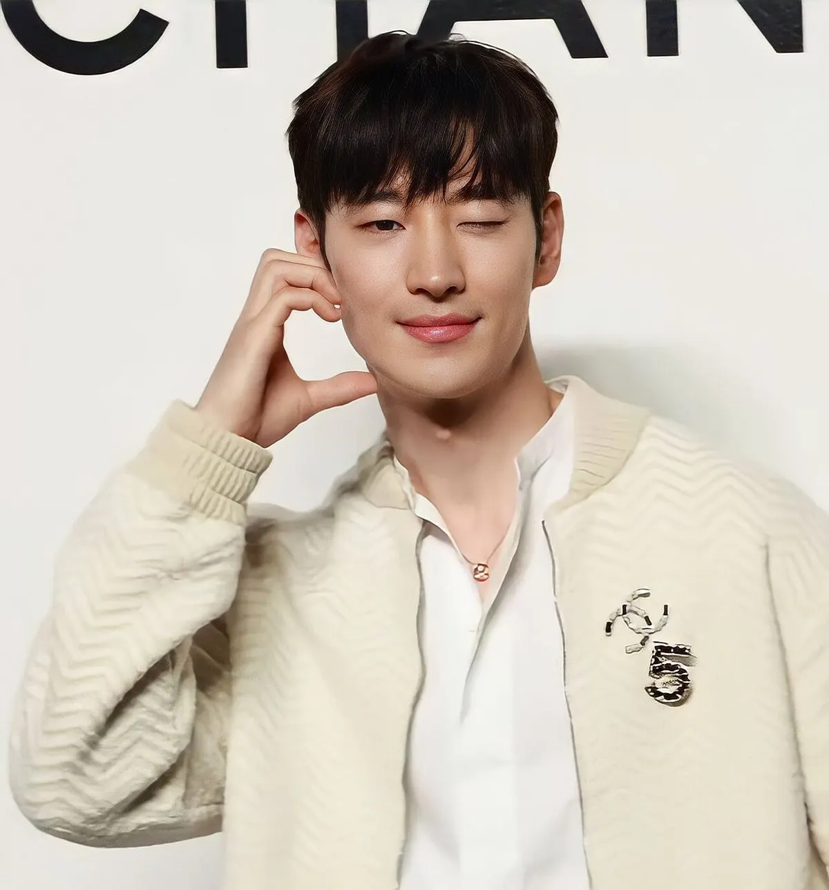 Lee Je-hoon berjaket krem bermotif zigzag tersenyum sambil mengedipkan satu mata di depan dinding putih bertulisan hitam