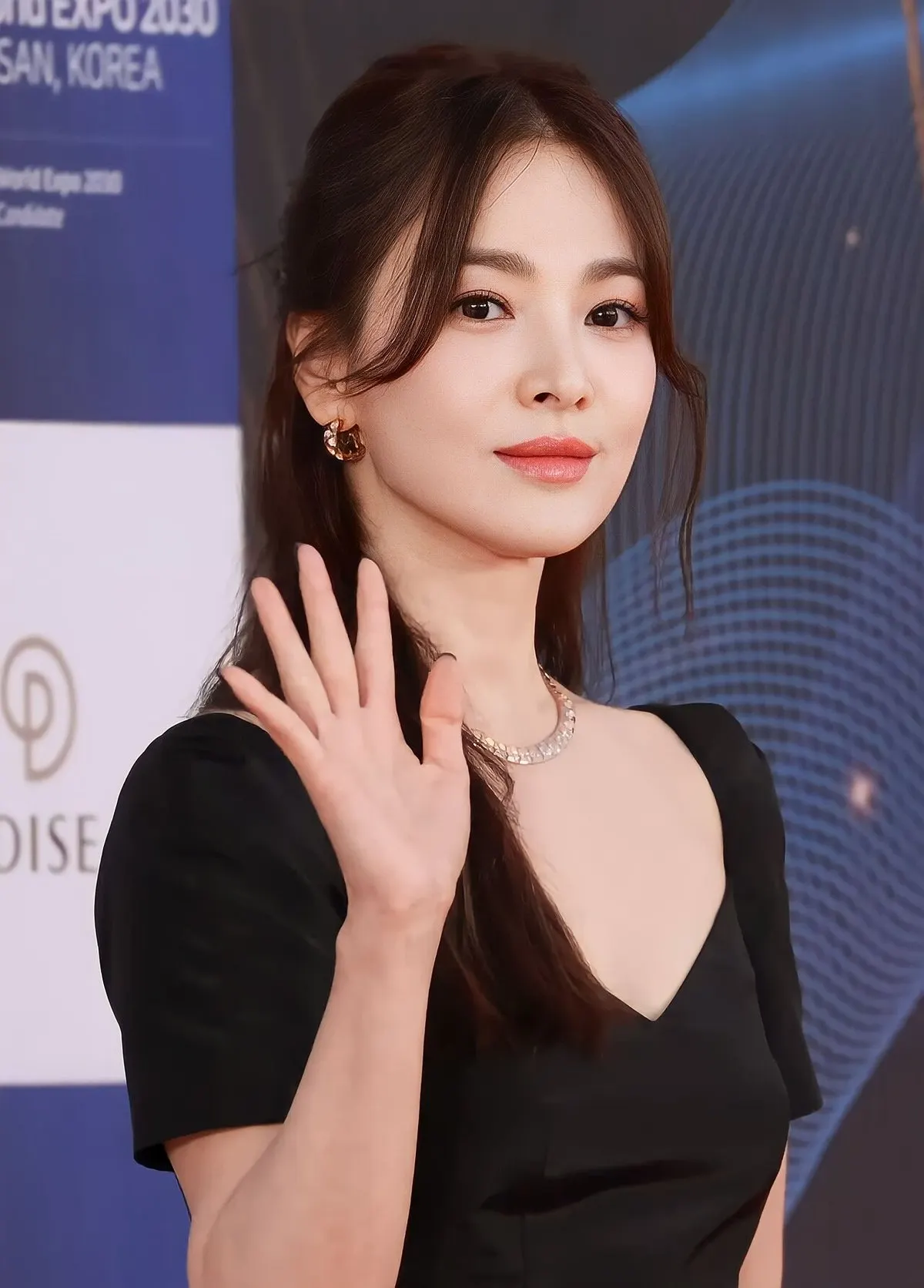 Song Hye-kyo bergaun hitam dan berkalung perak melambaikan tangan di karpet merah dengan latar papan acara berwarna biru