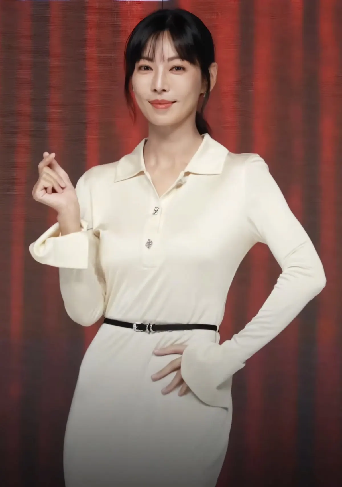 Aktris Kim So-yeon berpose dengan gestur hati jari, mengenakan atasan krem berkerah dan ikat pinggang hitam di depan latar merah