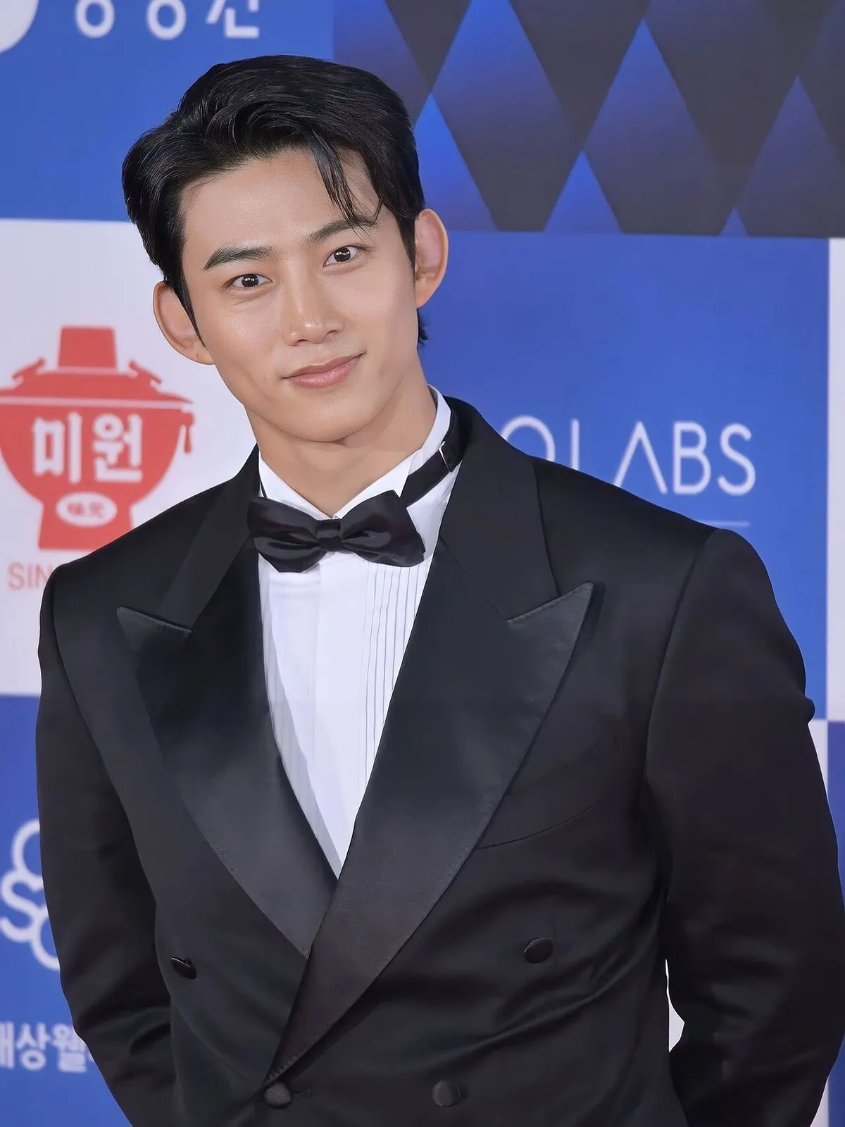 Aktor Ok Taec-yeon tersenyum mengenakan tuksedo hitam dan dasi kupu-kupu di depan papan sponsor berwarna biru