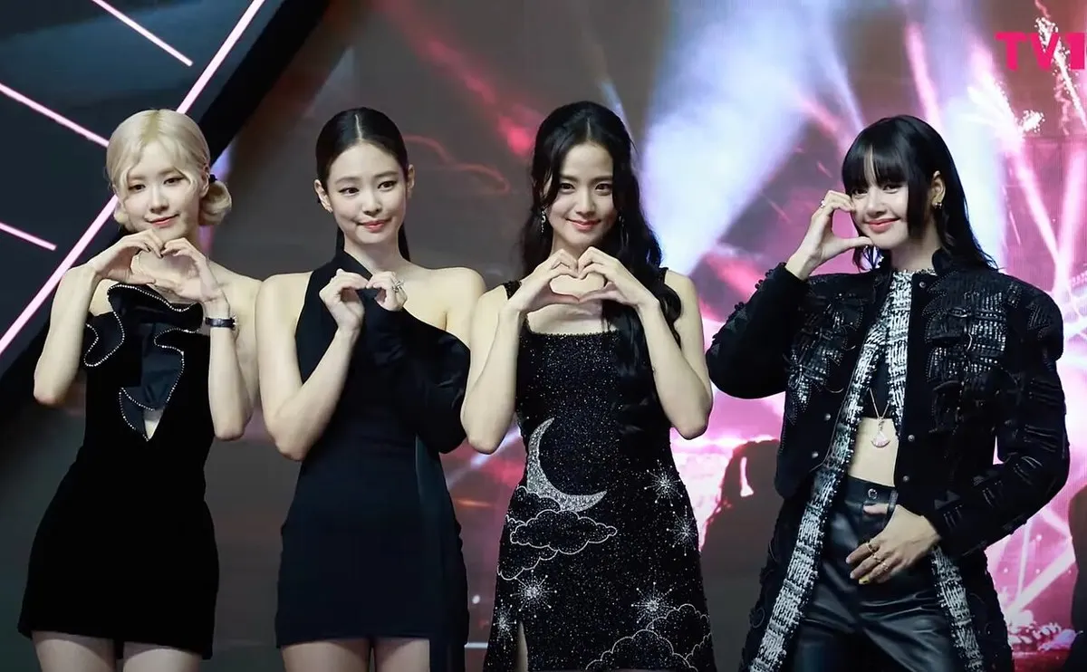 Empat anggota BLACKPINK berpose membentuk hati dengan tangan di depan latar panggung berlampu merah muda