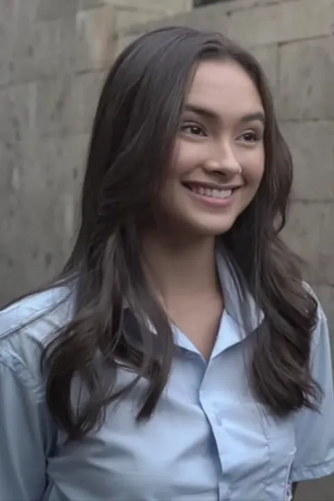 Caitlin Halderman berkemeja biru muda tersenyum di depan dinding batu saat produksi serial