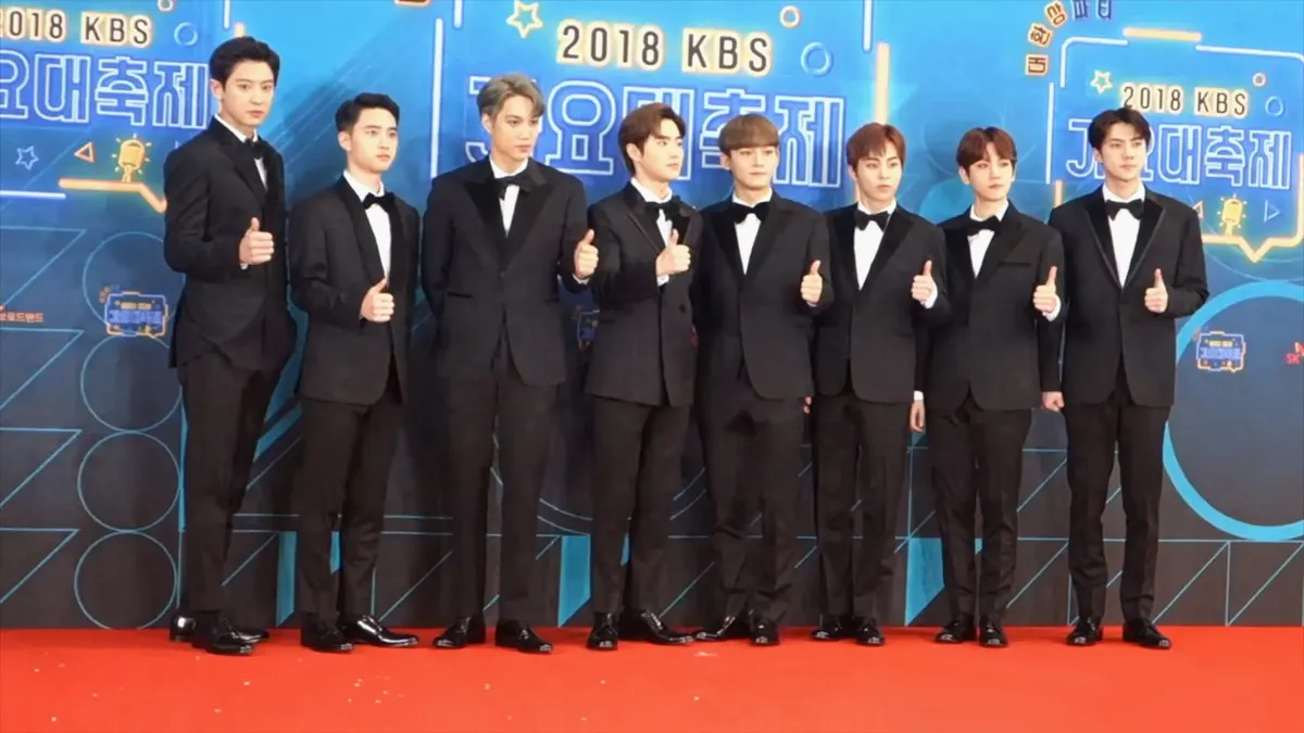 Anggota EXO berjas hitam berdiri berjajar di karpet merah dengan latar logo KBS