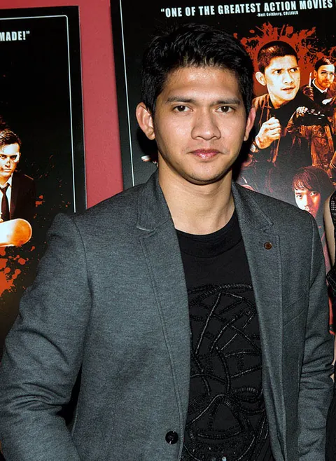 Iko Uwais berblazer abu-abu dan kaus hitam berpose di depan poster film laga