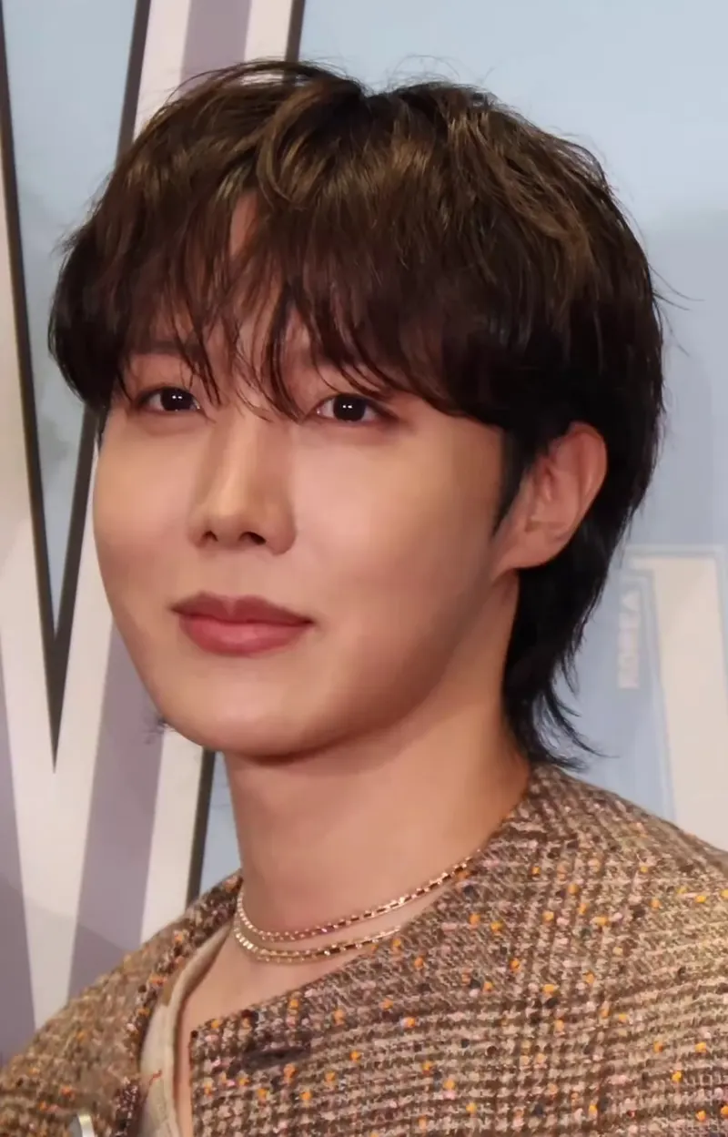 j-hope berambut ikal dengan jas bermotif dan kalung tipis berpose ke kamera