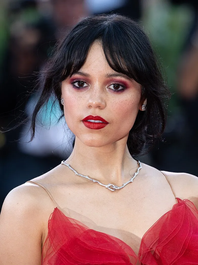 Jenna Ortega bergaun merah dan berkalung perak dengan latar belakang buram