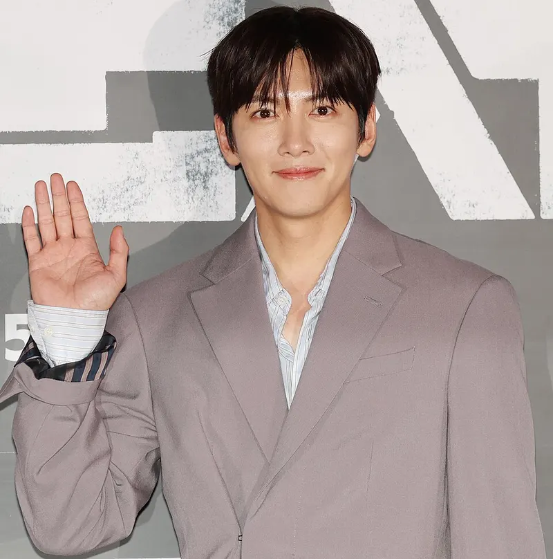 Ji Chang-wook berjas abu-abu kecokelatan melambaikan tangan kanan di depan latar poster