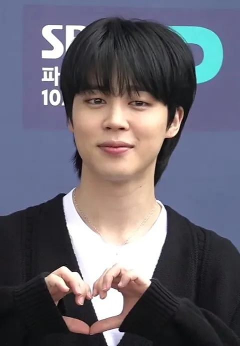 Jimin bersweter hitam membentuk hati dengan kedua tangan di depan papan SBS
