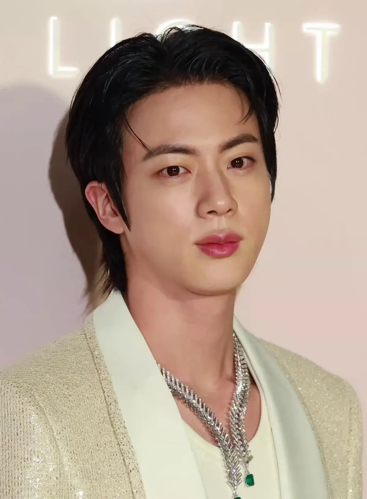 Jin berjas krem dengan kalung perak berpose di depan dinding bertuliskan LIGHT