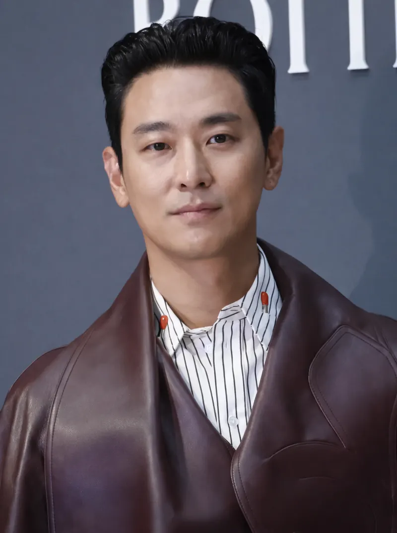 Ju Ji-hoon bermantel kulit merah marun dan kemeja bergaris di depan latar biru keabuan