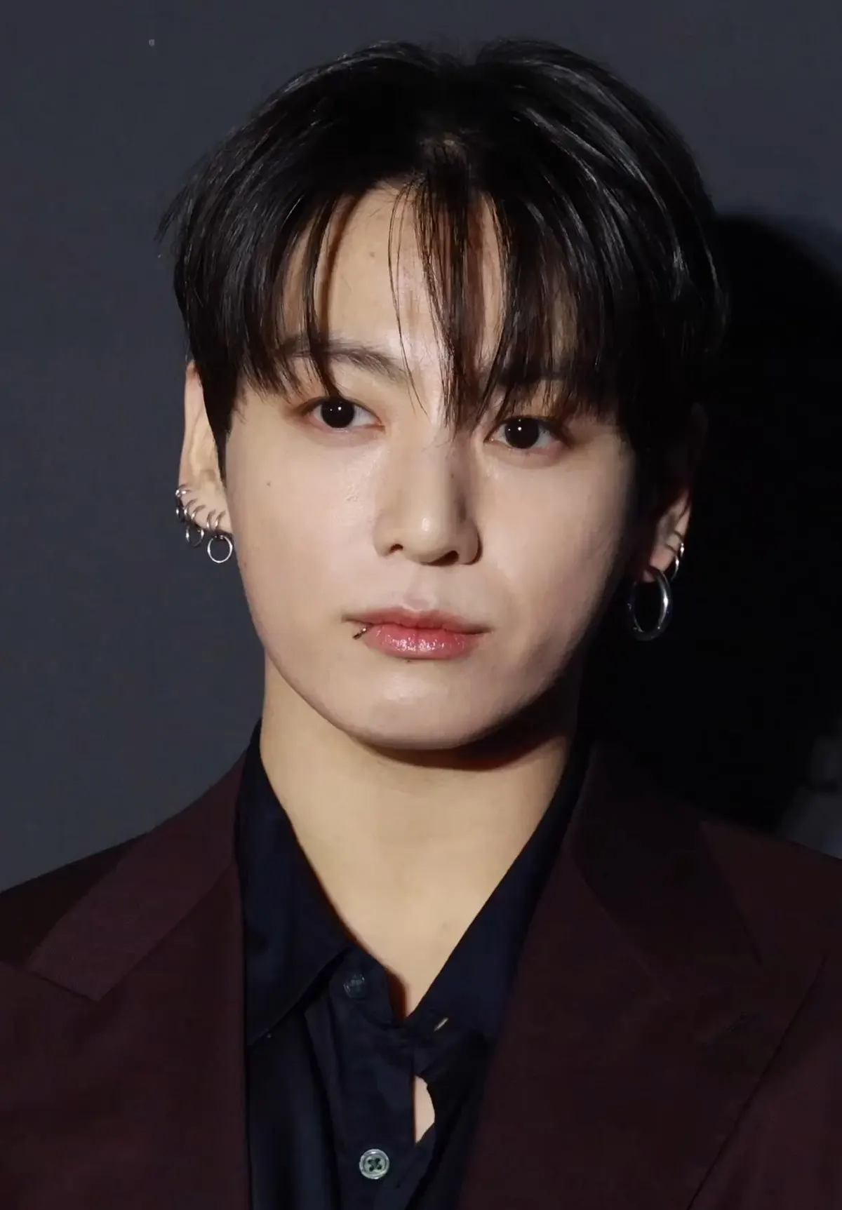 Jungkook berjas merah marun dengan anting perak menatap kamera di depan latar gelap