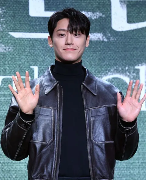 Lee Do-hyun berjaket kulit hitam dan turtleneck melambaikan kedua tangan di jumpa pers