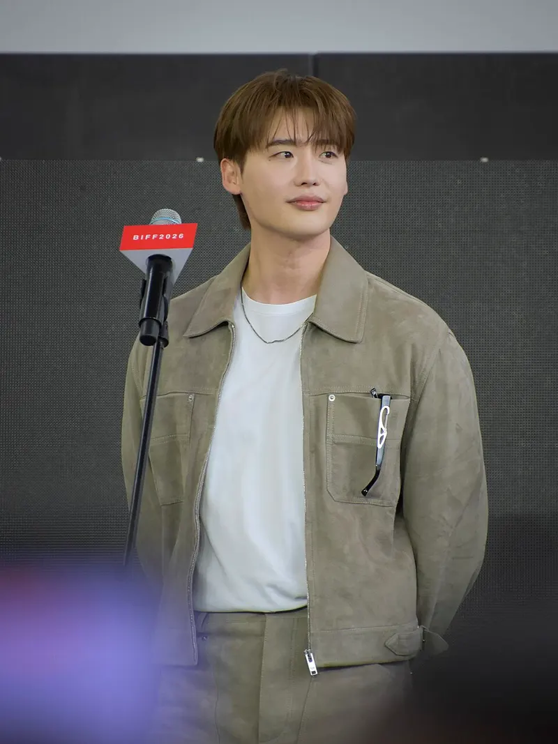 Lee Jong-suk berjaket suede krem berdiri di belakang mikrofon berlabel BIFF2026
