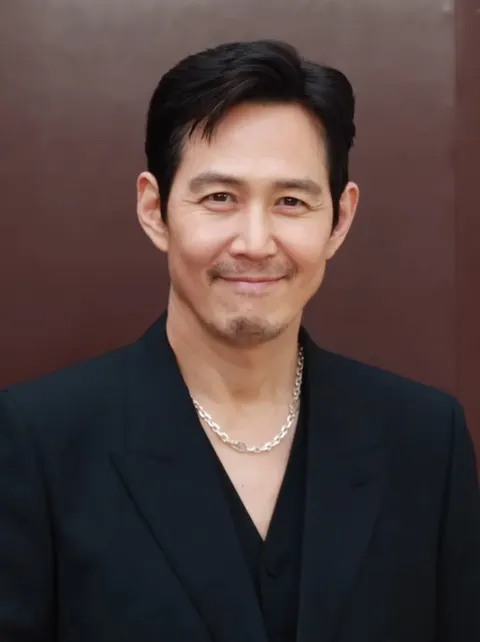 Lee Jung-jae berjas hitam dan berkalung rantai perak tersenyum di depan latar cokelat