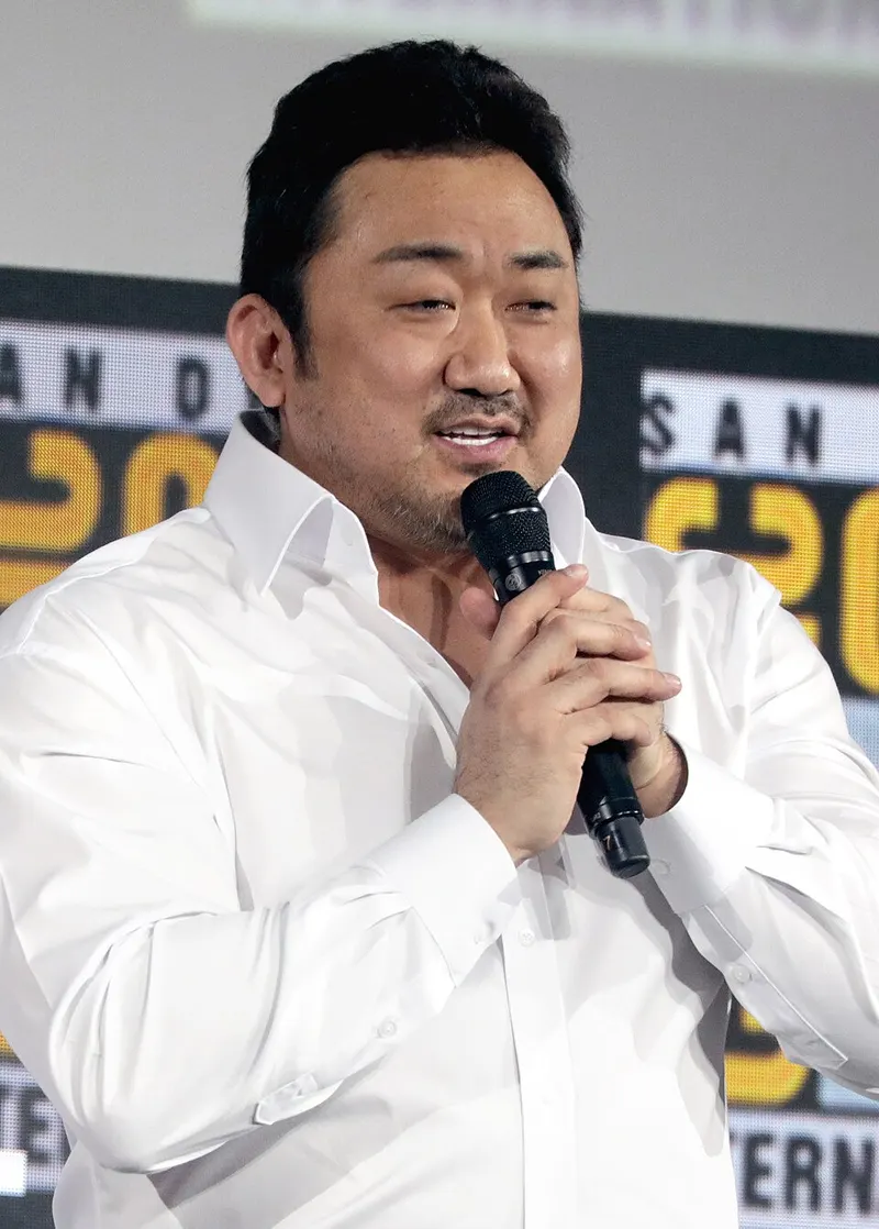 Ma Dong-seok berkemeja putih berbicara sambil memegang mikrofon di panggung