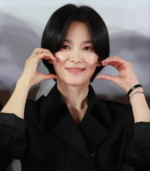 Song Hye-kyo berambut pendek dan bermantel hitam, kedua tangan diangkat di dekat wajah