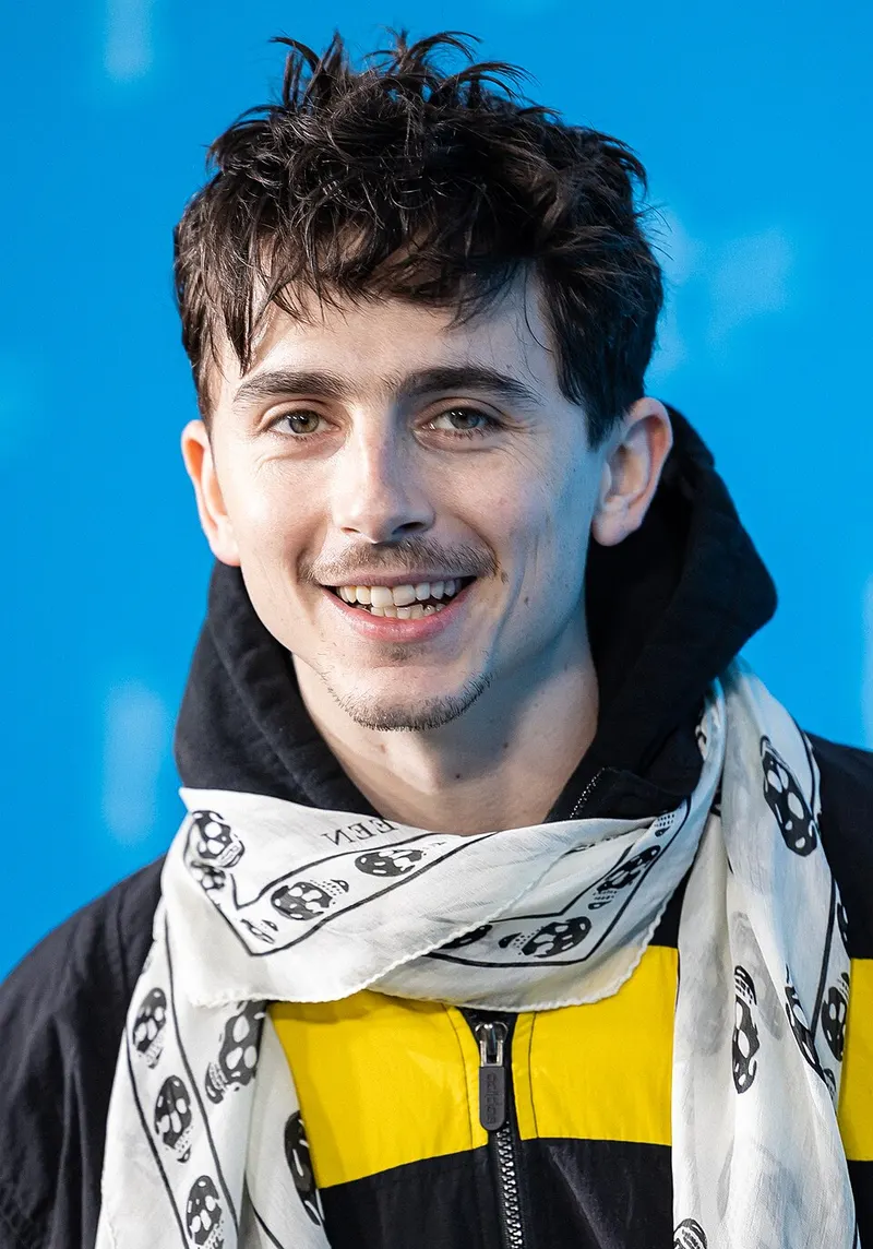 Timothée Chalamet berjaket hitam kuning dan berselendang putih bermotif tengkorak tersenyum