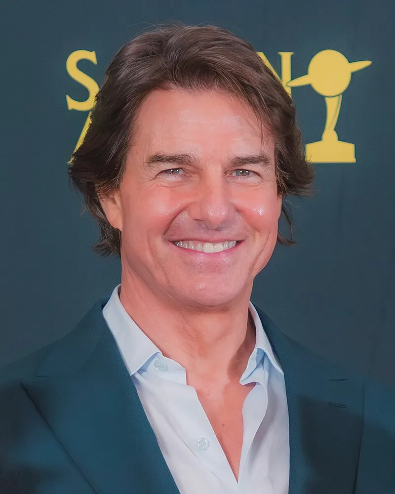 Tom Cruise berjas biru tua dan berkemeja putih tersenyum di depan papan Saturn Awards