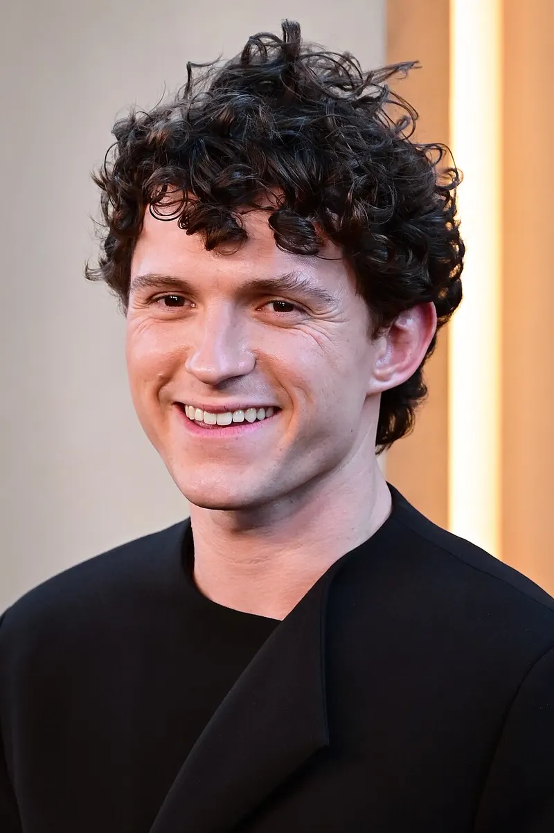 Tom Holland berambut ikal dan berbaju hitam tersenyum di sebuah acara