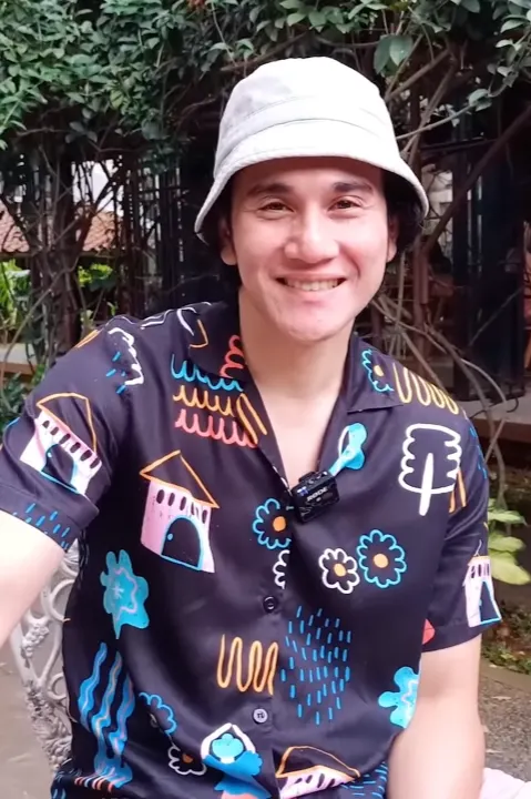 Vino G. Bastian bertopi bucket putih dan berkemeja motif tersenyum di taman